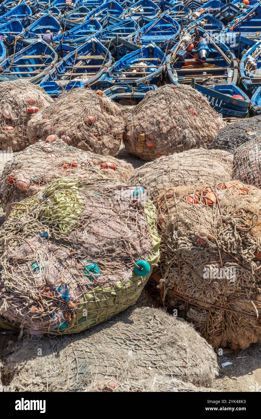 Reti da pesca raccolte in grandi balle su una banchina nel porto peschereccio di Essaouira in Marocco. Una flottiglia di piccole barche da pesca sullo sfondo Foto Stock