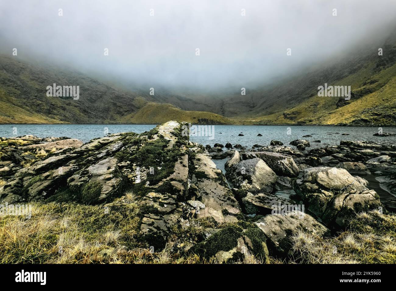 Un corpo d'acqua incastonato tra una catena montuosa nebbiosa. L'acqua è calma e riflette il cielo nuvoloso. Rocce e erba sono sparse lungo la riva. Foto Stock