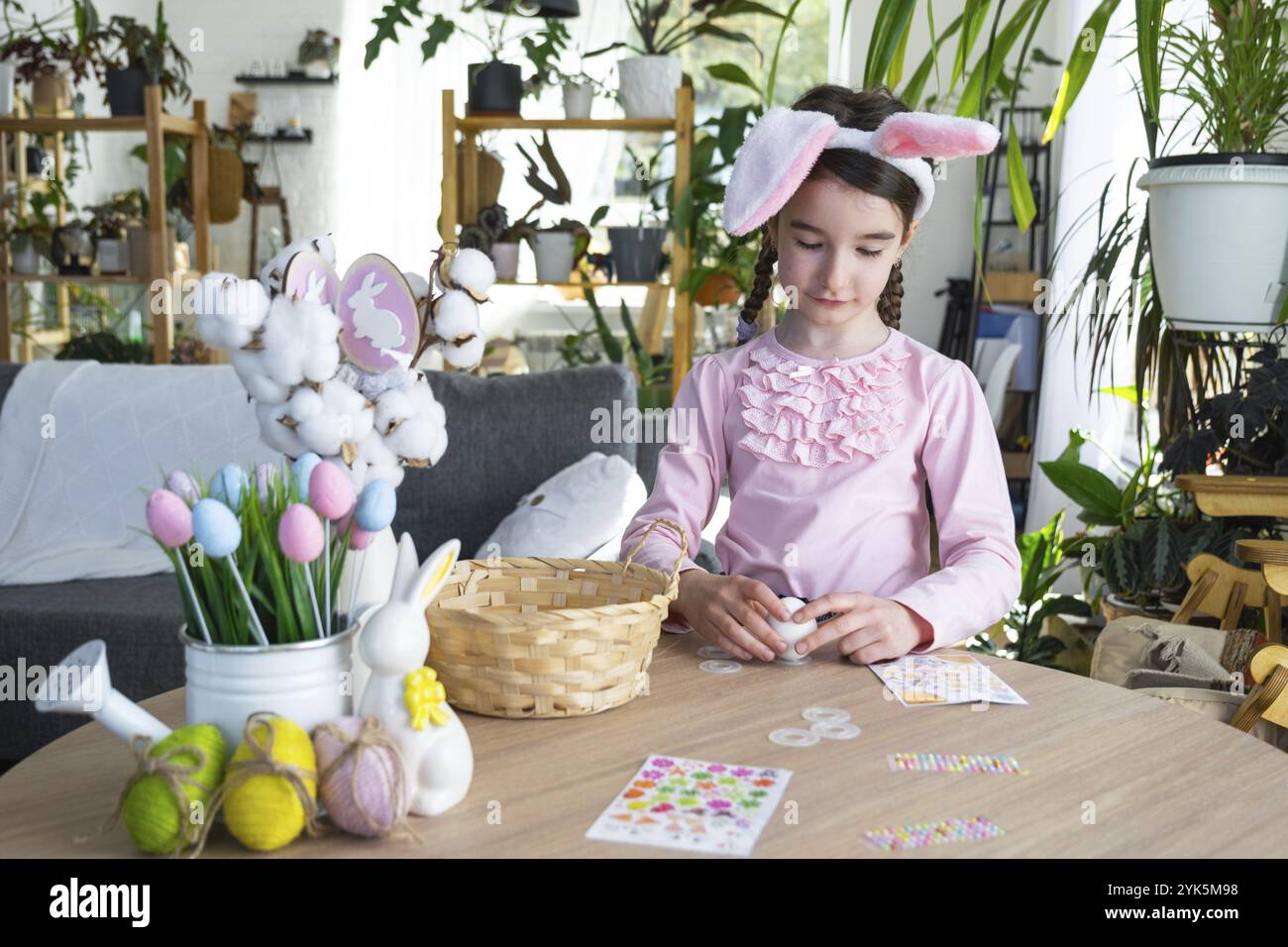 Una ragazza carina con le orecchie rosa coniglietto crea un artigianato pasquale, decora un uovo a forma di unicorno con strass, corno, fiori all'interno di un Foto Stock