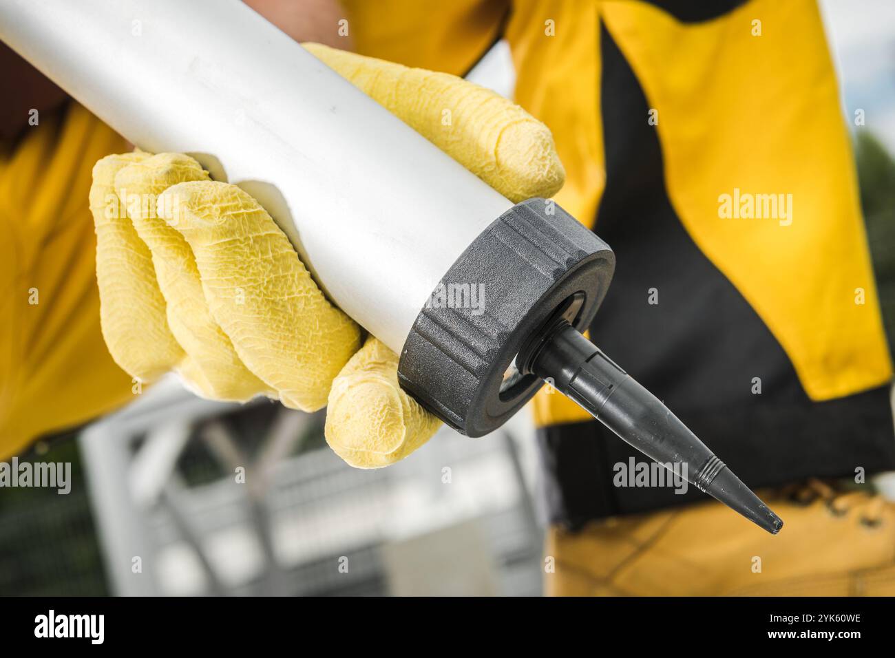 Un lavoratore afferra una pistola cianfrinante con guanti gialli, preparandosi per un'attività di manutenzione all'aperto, sotto il cielo limpido. L'impostazione è luminosa e ben illuminata. Foto Stock