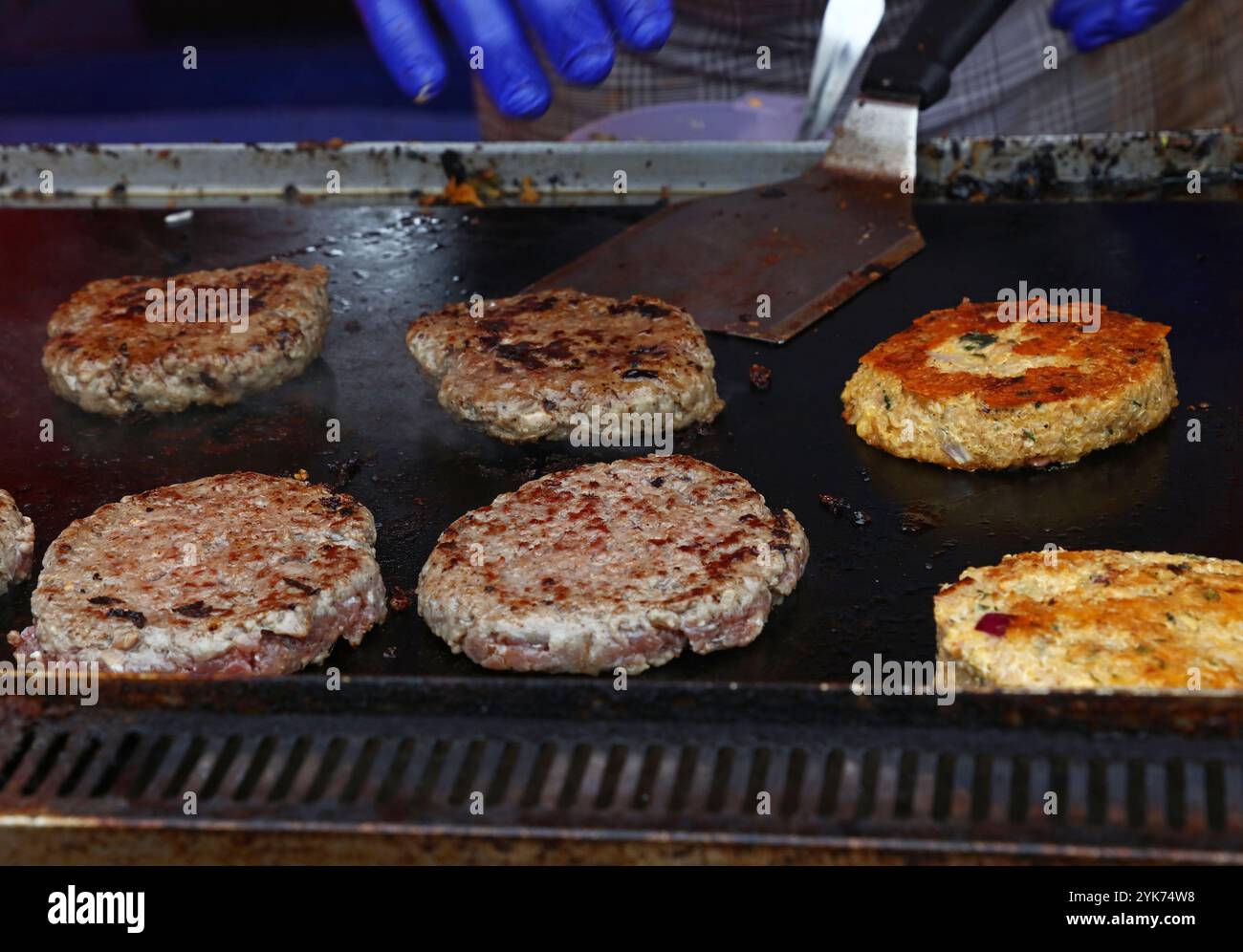 Cuocere con i guanti deliziosi hamburger e hamburger vegetariani su una griglia calda, utilizzando una spatola per cuocerli in modo uniforme Foto Stock