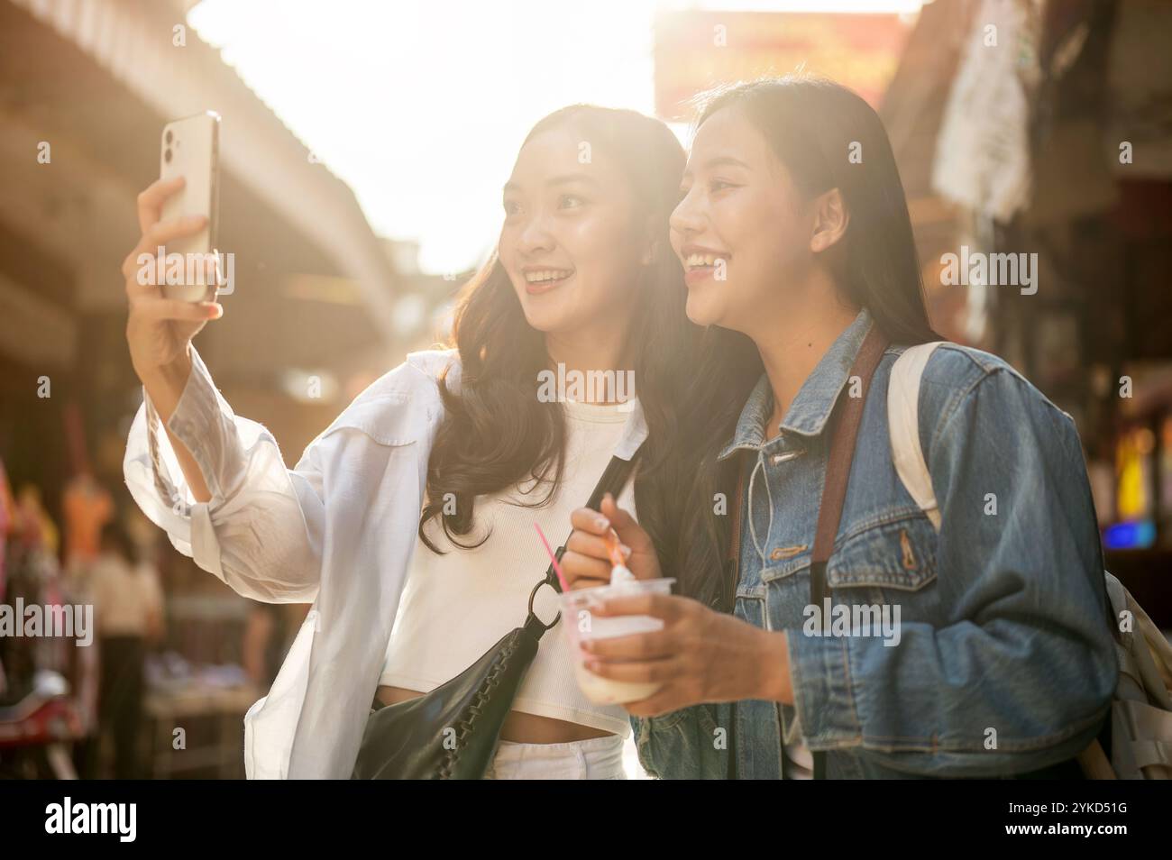 Due graziose e gioiose giovani turisti asiatiche amano esplorare il cibo di strada in un mercato in Thailandia in una giornata di sole, facendo un selfie insieme a un sma Foto Stock