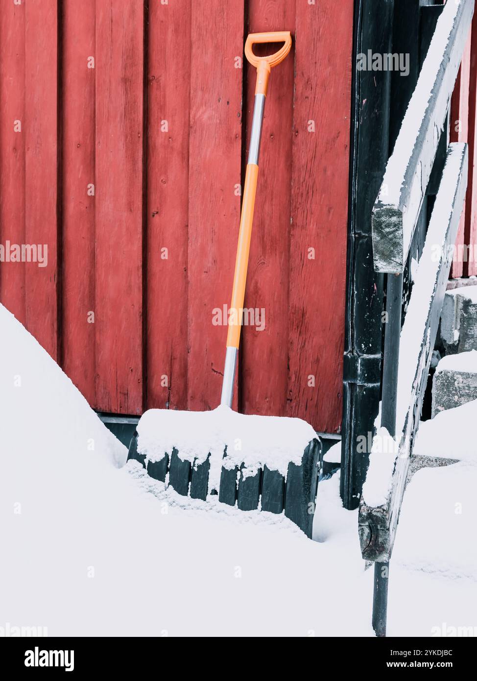 Una pala poggia contro un muro rosso, parzialmente sepolto nella neve. La scena raffigura una tipica giornata invernale, con neve bianca fresca che copre il terreno e il fiume Foto Stock