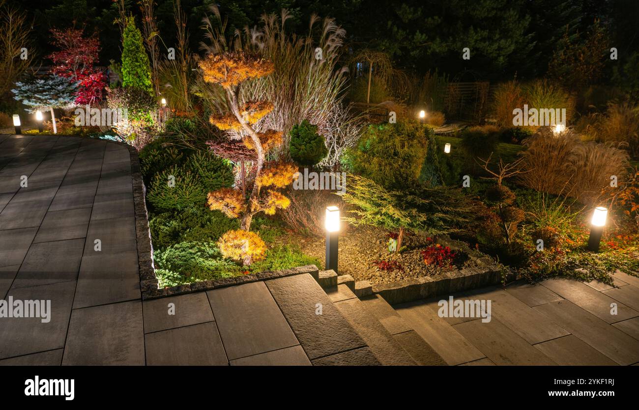 Luci calde illuminano il sentiero del giardino, mettendo in risalto alberi vibranti e piante curate, creando un'atmosfera serena per passeggiate serali. Foto Stock