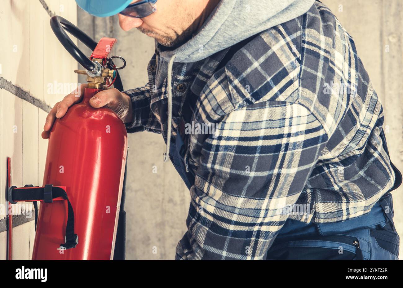 Un lavoratore vestito con una camicia a quadri ispeziona un estintore montato su una parete, garantendo la conformità alla sicurezza in un ambiente commerciale. Foto Stock