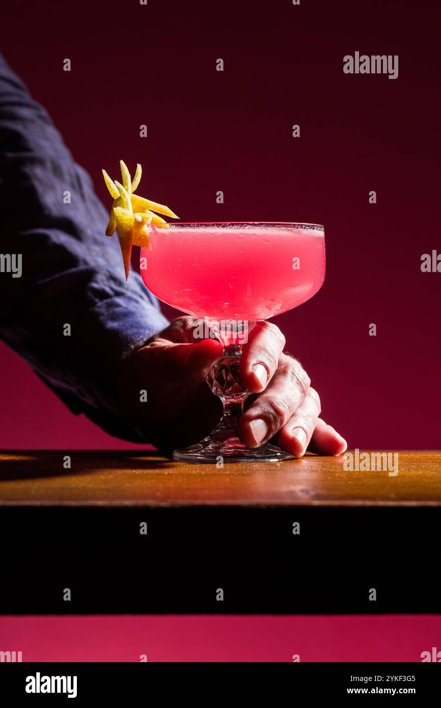 Il cocktail cosmopolita è presentato elegantemente in un bicchiere da coupé, adornato con un tocco di limone, la bevanda rosa contrasta splendidamente con il dorso Foto Stock