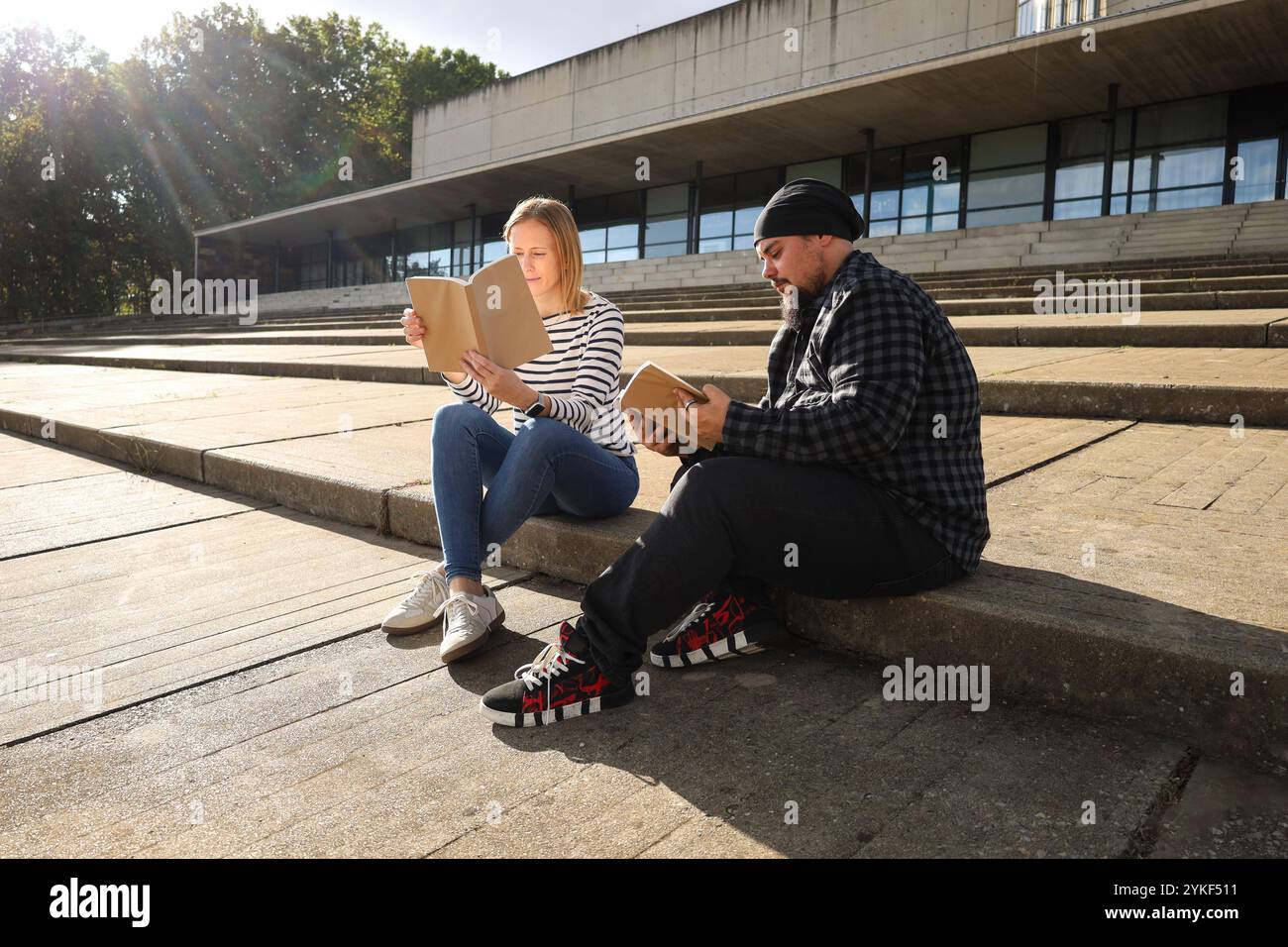 Un uomo trans e il suo amico si godono una giornata di sole leggendo libri sui gradini di un edificio moderno, mettendo in evidenza temi di amicizia e orgoglio LGBTQIA+ Foto Stock