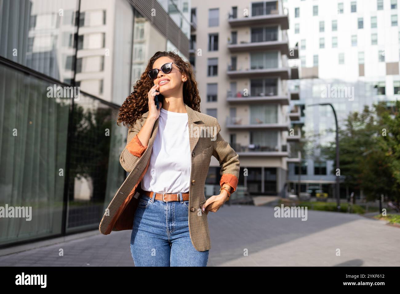 Un'elegante donna d'affari brasiliana cammina con sicurezza mentre parla sul suo smartphone fuori dagli edifici moderni è vestita con un blazer chic e una sungla Foto Stock