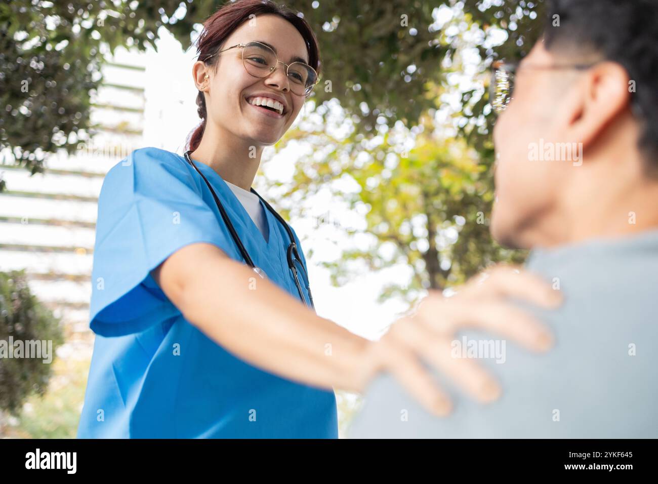 Una giovane studentessa infermieristica, vestita con un abito blu e occhiali, sorride mentre aiuta un paziente all'aperto, aiutandolo, circondato dal verde Foto Stock