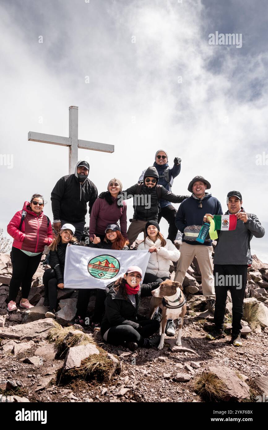 Un vivace gruppo di escursionisti raggiunge una cima di montagna, potendo con gioia due bandiere da una grande croce, accanto a un cane amico, aggiunge Un cielo nuvoloso Foto Stock