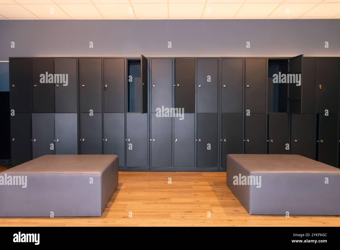 Vista interna di uno spogliatoio vuoto della palestra con file di armadietti grigi e due eleganti panche su un pavimento in legno. Foto Stock