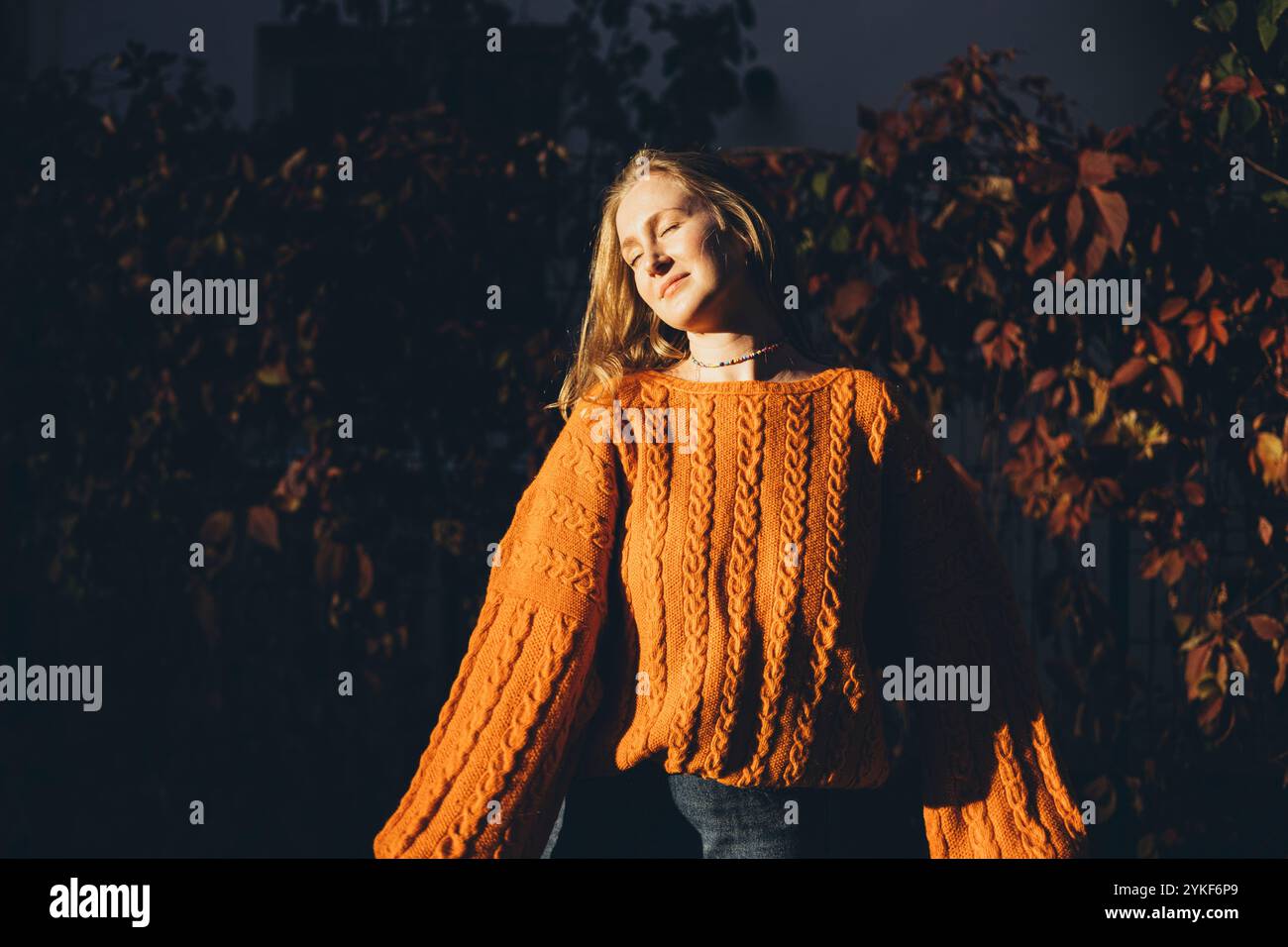 Una donna si crogiola al calore della luce del sole, indossando un comodo maglione in maglia arancione le tonalità calde mettono in risalto l'ambiente autunnale, con uno sfondo di foglie Foto Stock