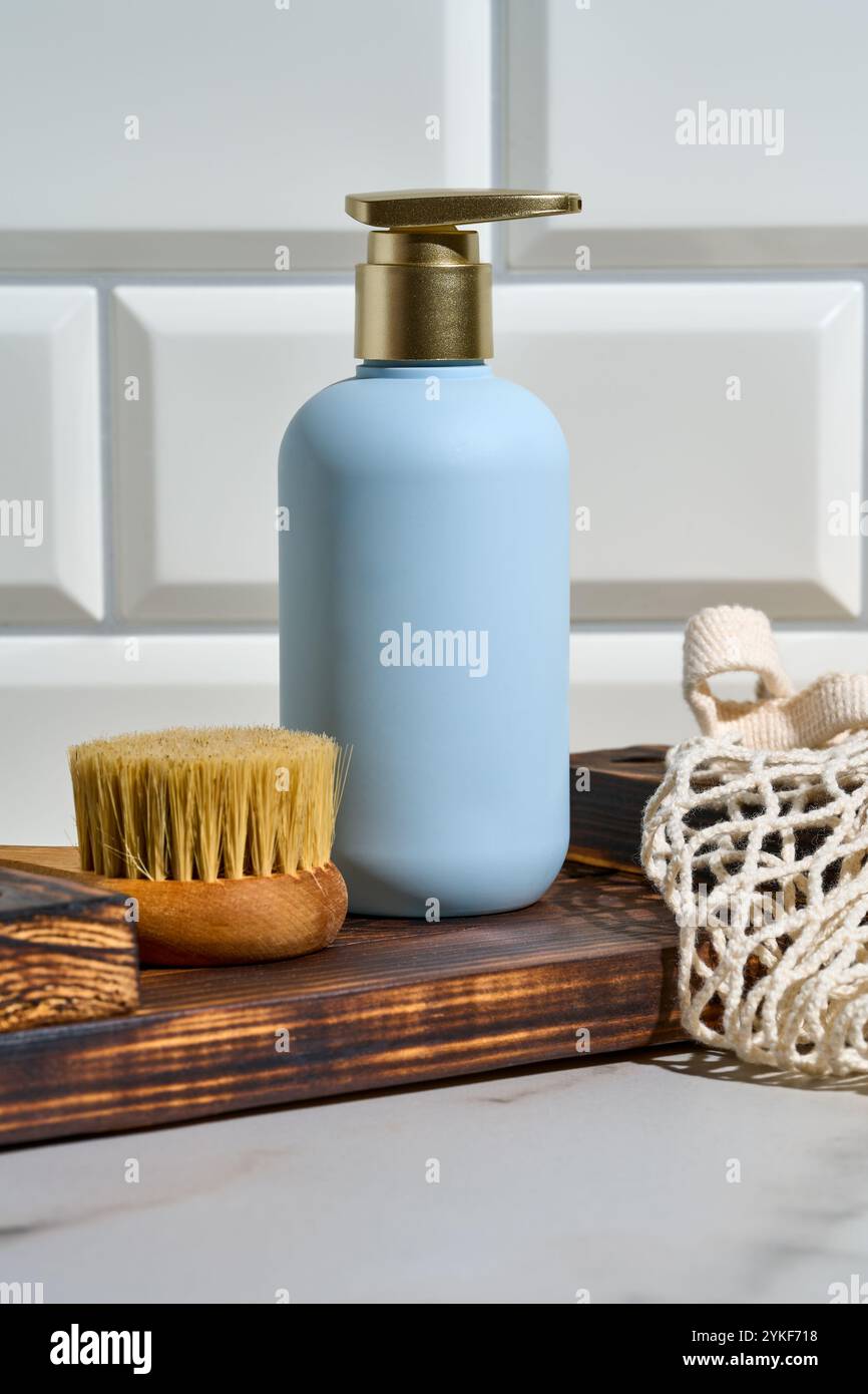 Un'elegante bottiglia cosmetica SPA blu opaco con dispenser a tiralatte dorato si trova su un ripiano in legno accanto alle spazzole a setole naturali e a una rete, su una ba luminosa Foto Stock