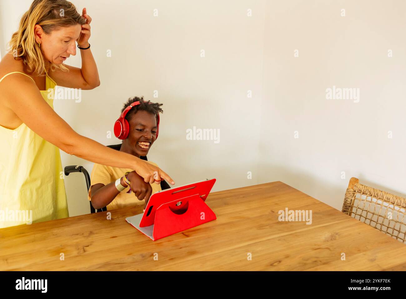 Un giovane ragazzo nero con paralisi cerebrale interagisce gioiosamente con la sua insegnante femminile in una casa di vacanza. Utilizza un tablet e indossa le cuffie mentre lei lo fa Foto Stock