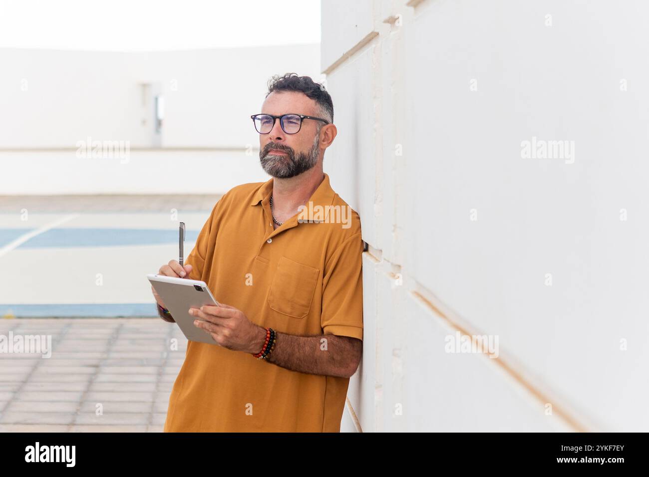 Un uomo gay di mezza età con barba, occhiali e camicia casual si trova vicino a una parete esterna luminosa, tenendo in mano un tablet e una penna digitale, Foto Stock