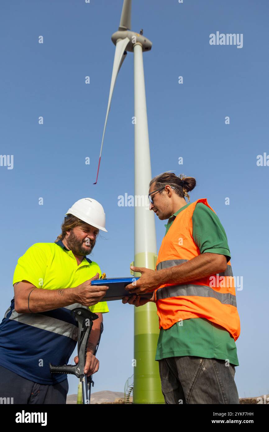 Due uomini, uno disabile, in attrezzatura di sicurezza discutono la sicurezza operativa di fronte a una torreggiante turbina eolica, evidenziando l'inclusività nella sfera delle energie rinnovabili Foto Stock