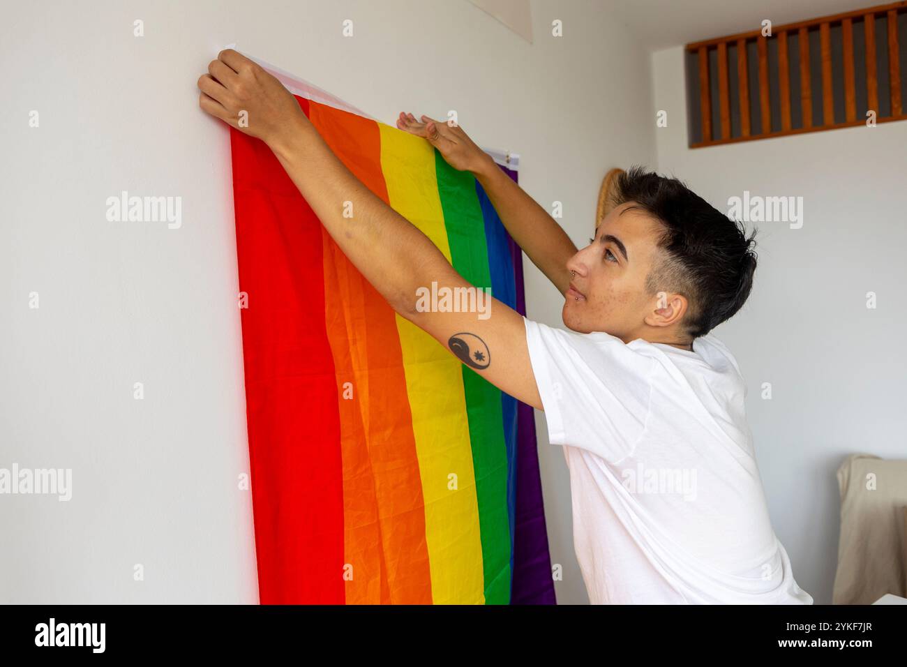 Un giovane trans appende una colorata bandiera d'orgoglio sul suo muro bianco, decorando la sua nuova casa ed esprimendo la sua identità Foto Stock