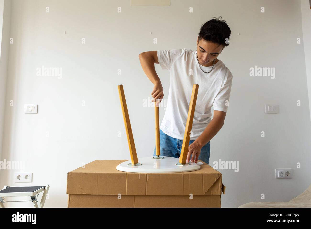 Un giovane trans è impegnato a assemblare un tavolo di legno nel suo luminoso e nuovo appartamento, simboleggiando un nuovo inizio e l'autosufficienza Foto Stock