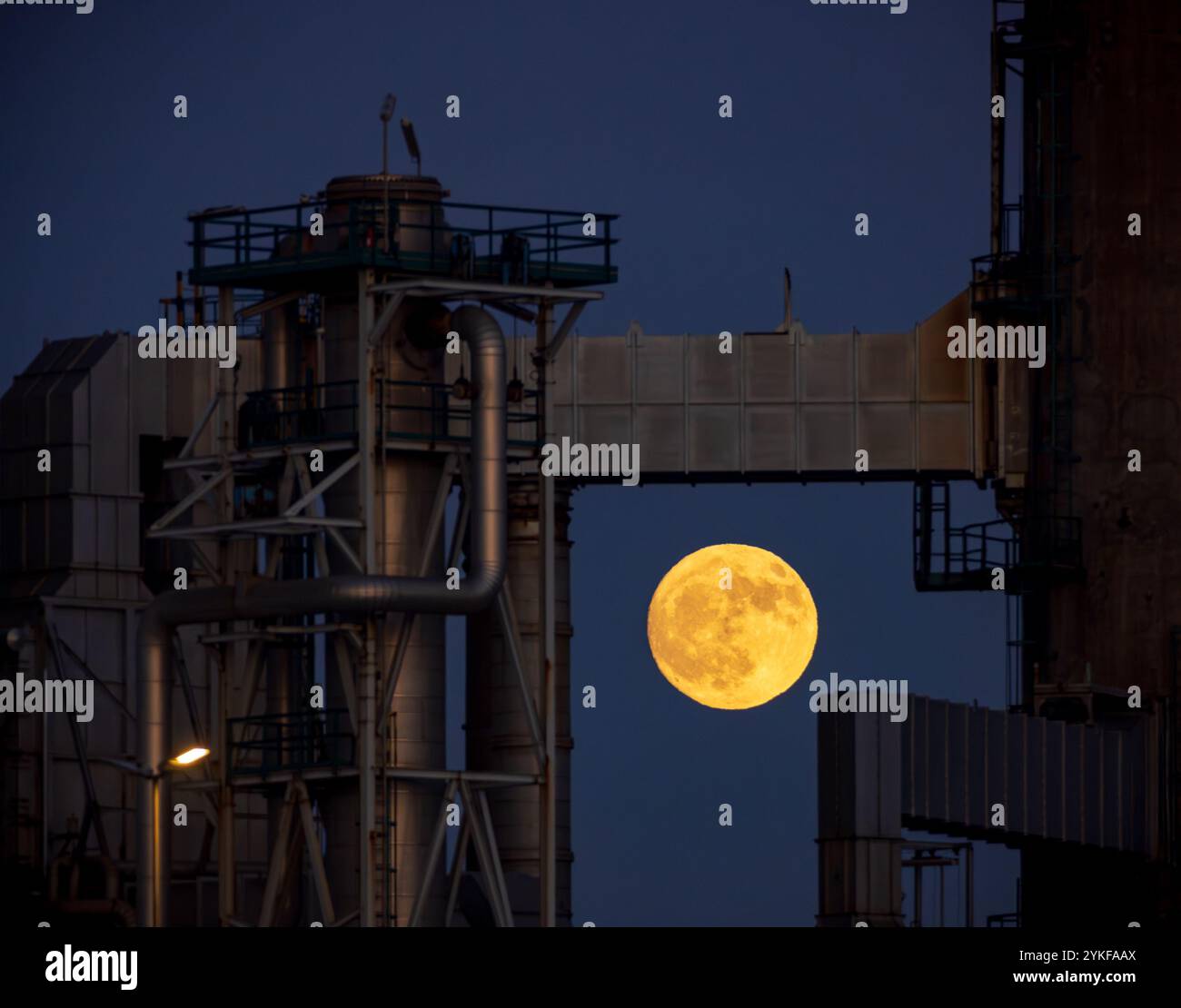 Un'impressionante luna piena sale nel cielo blu scuro dietro la silhouette di complesse installazioni industriali a Porto, Portogallo. Foto Stock