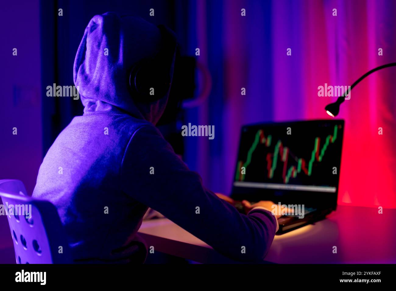 Vista posteriore di un ragazzo irriconoscibile che commercializza azioni su un notebook, evidenziata da una luce colorata dello schermo in una stanza buia. L'immagine cattura un momento di pausa Foto Stock