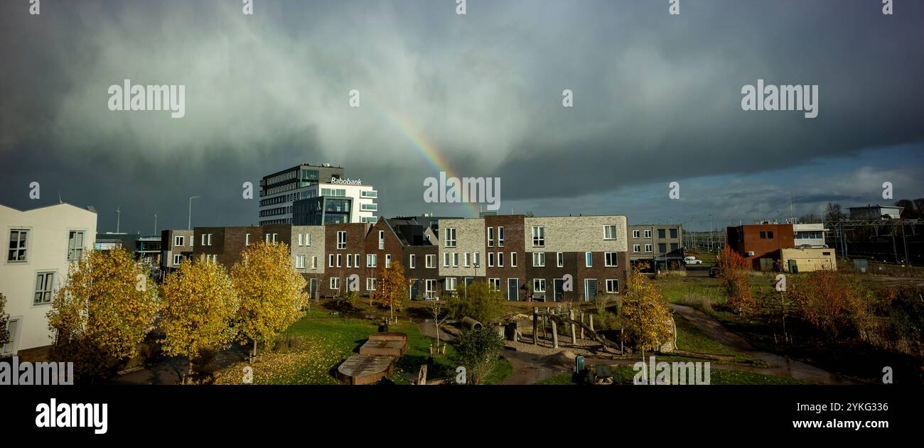 Arcobaleno e nuvole di tempesta piovosa sopra la banca dei contadini olandesi Rabobank e il parco giochi del quartiere autunnale Ettegerpark in primo piano illuminato dal sole Foto Stock