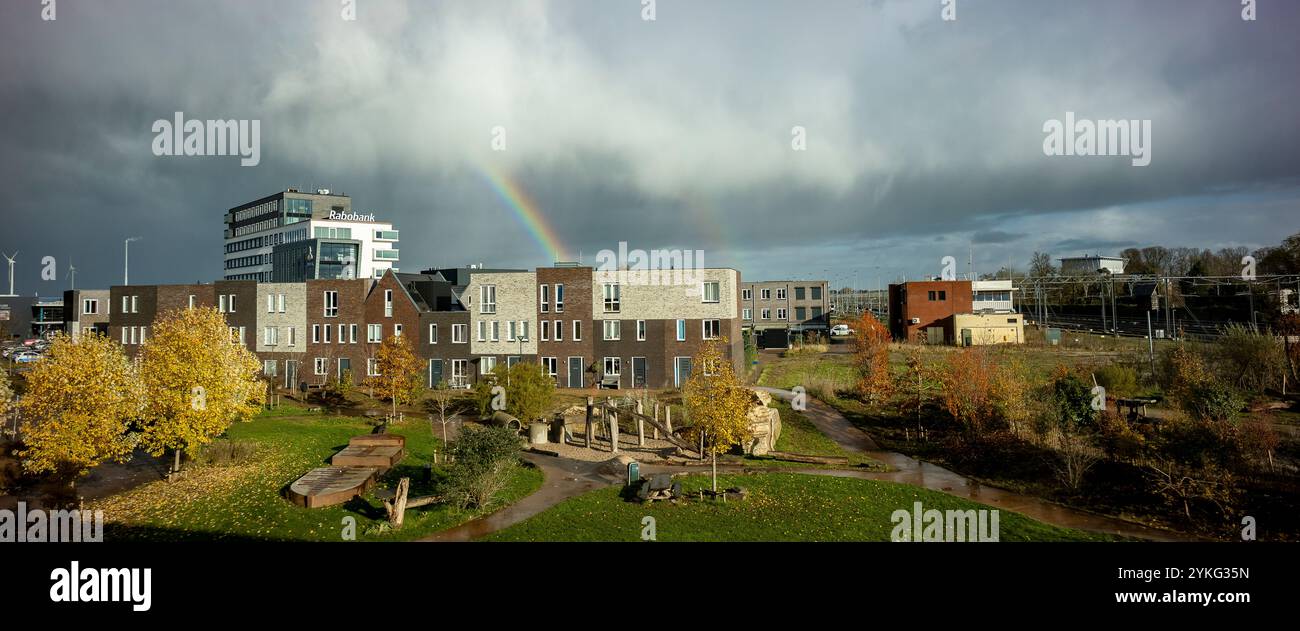 Arcobaleno e nuvole di tempesta piovosa sopra la banca dei contadini olandesi Rabobank e il parco giochi del quartiere autunnale Ettegerpark in primo piano illuminato dal sole Foto Stock