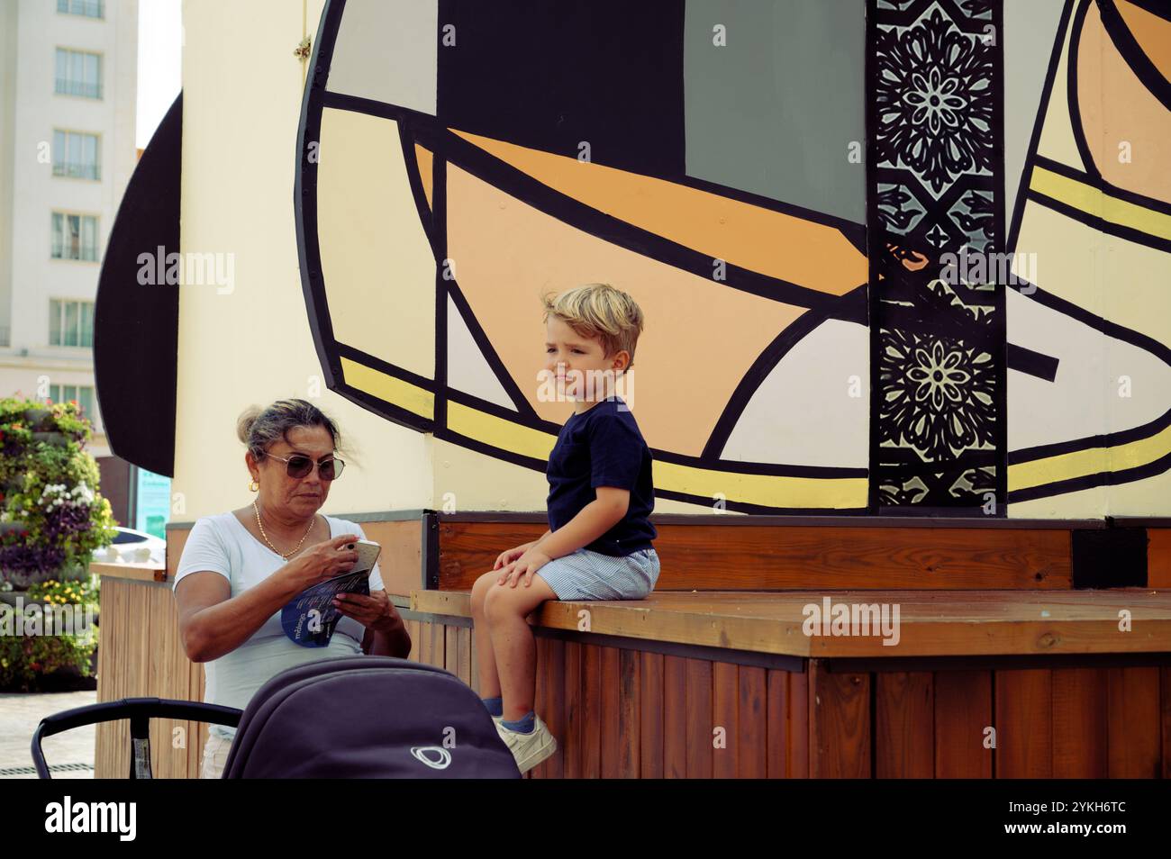 La babysitter controlla il suo telefono da bambino seduto su una piattaforma di legno su un colorato sfondo murale Foto Stock