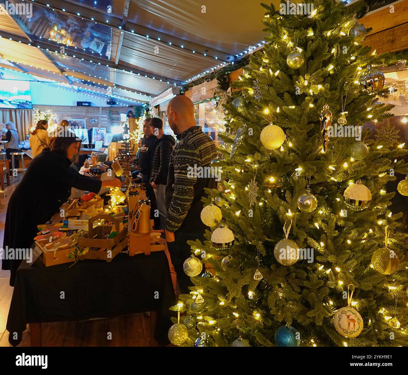 Gli amanti dello shopping e dei banchi che partecipano a una fiera di artigianato natalizio dominata da un albero di Natale decorato con colori vivaci Foto Stock