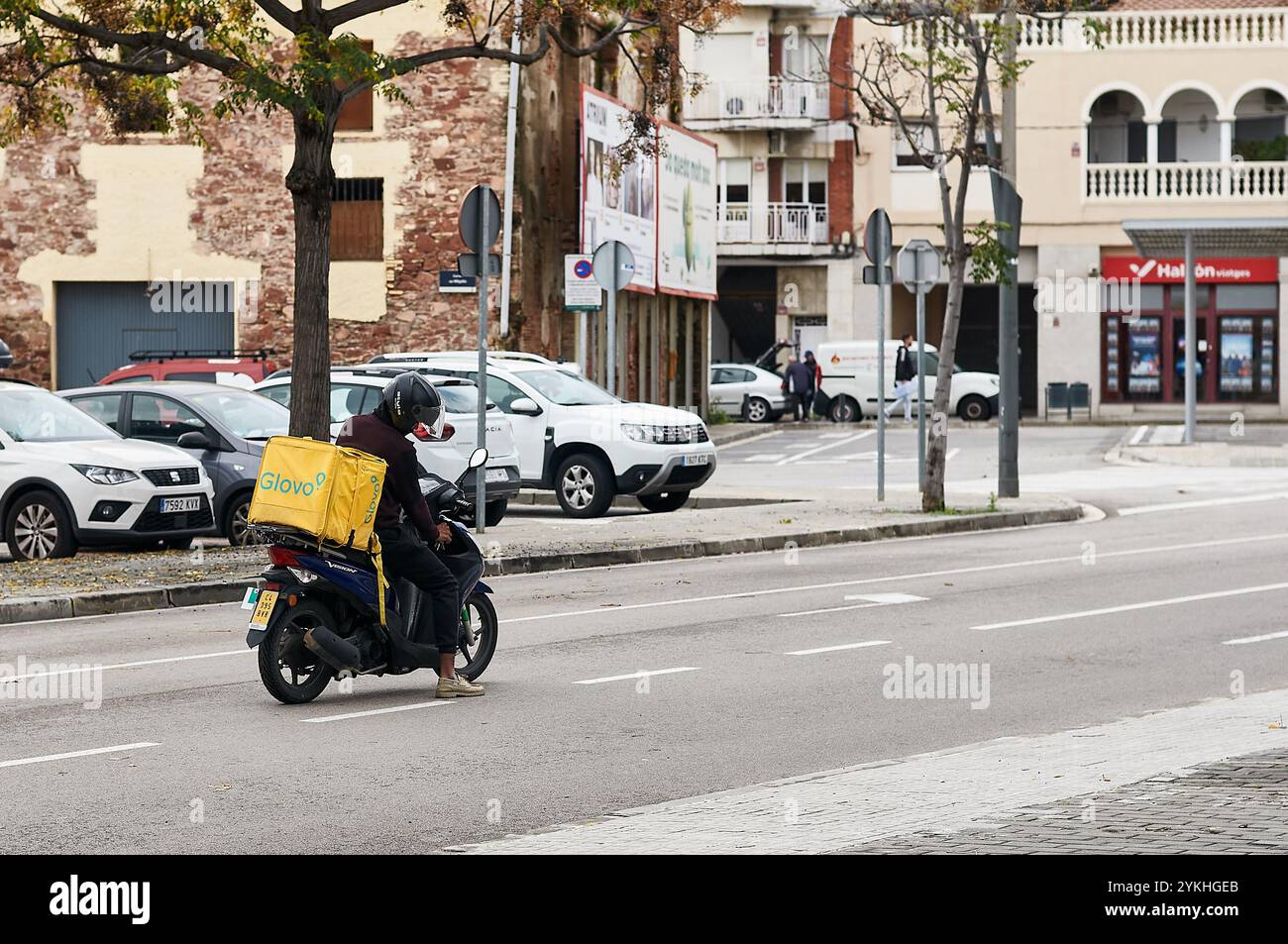 Viladecans. Spagna - 18 novembre 2024: Glovo corriere con la sua moto parcheggiata in mezzo alla strada e lo zaino per la consegna. La sc Foto Stock