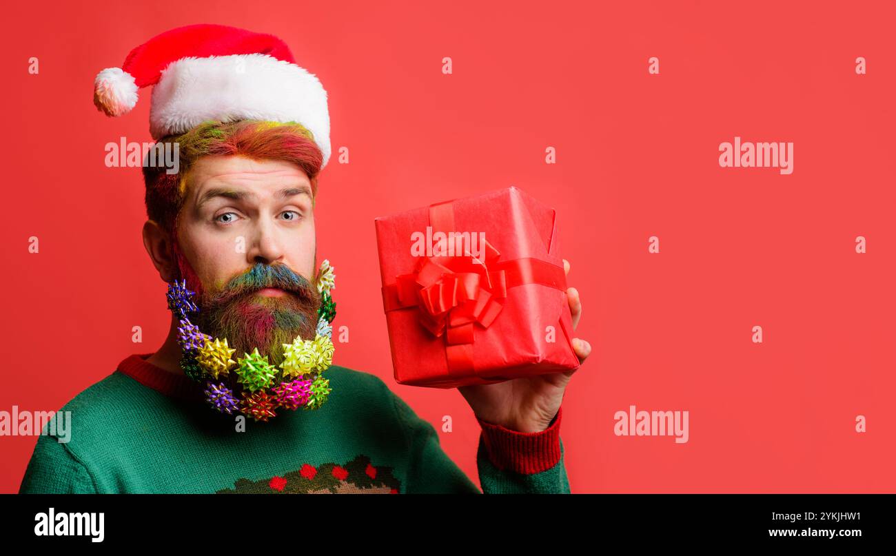 Shopping di Natale. Uomo serio con barba decorata in cappello di Babbo Natale con regalo di Natale. Babbo Natale con baubles di Natale in barba tinto in scatola regalo Foto Stock