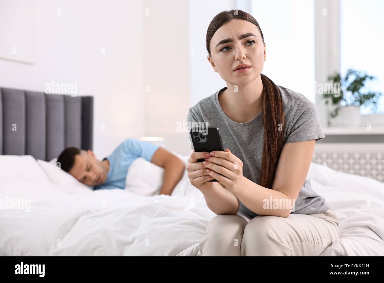 Donna frustrata con lo smartphone del marito mentre dorme in camera da letto, concentrazione selettiva Foto Stock