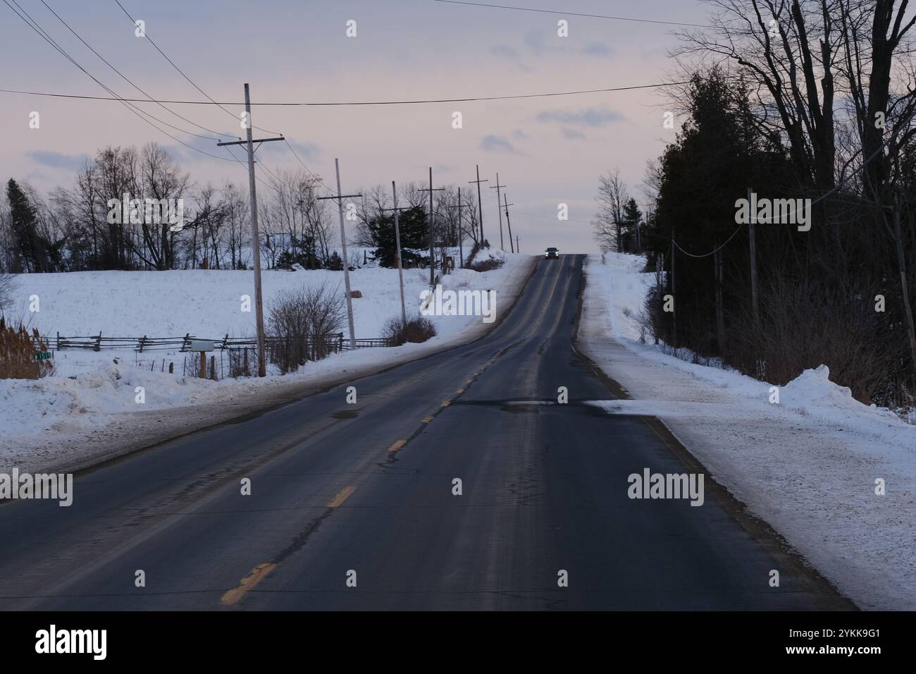 Paesaggi invernali e una strada con un po' di neve alla deriva. Regione dei laghi Kawartha, Ontario. Un'auto appare in cima alla collina. Foto Stock