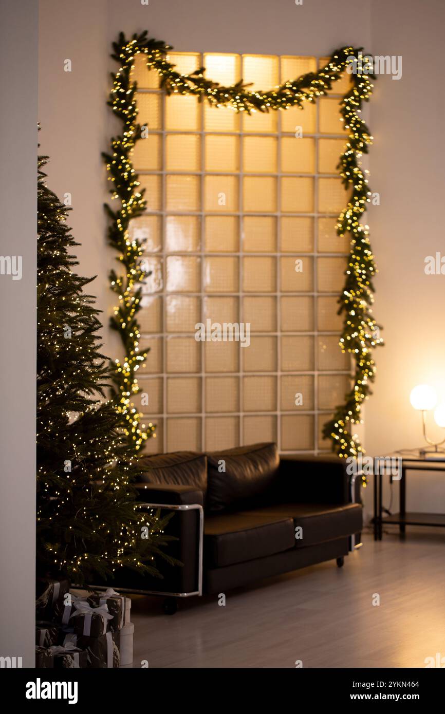 Davanti a un albero di Natale c'è un divano nero e una partizione fatta di blocchi di vetro. La camera è decorata con luci di Natale e un albero di Natale Foto Stock