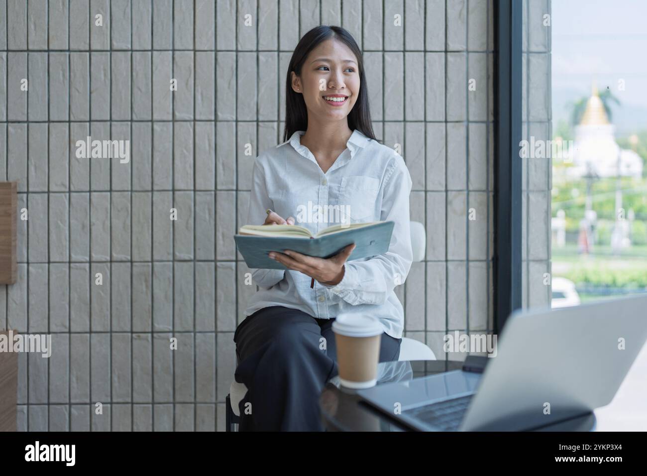 Una giovane donna gode di una sessione di studio produttiva in un bar moderno, sorridendo mentre prende appunti nel suo notebook, circondato da un portatile e caffè, incarnato Foto Stock