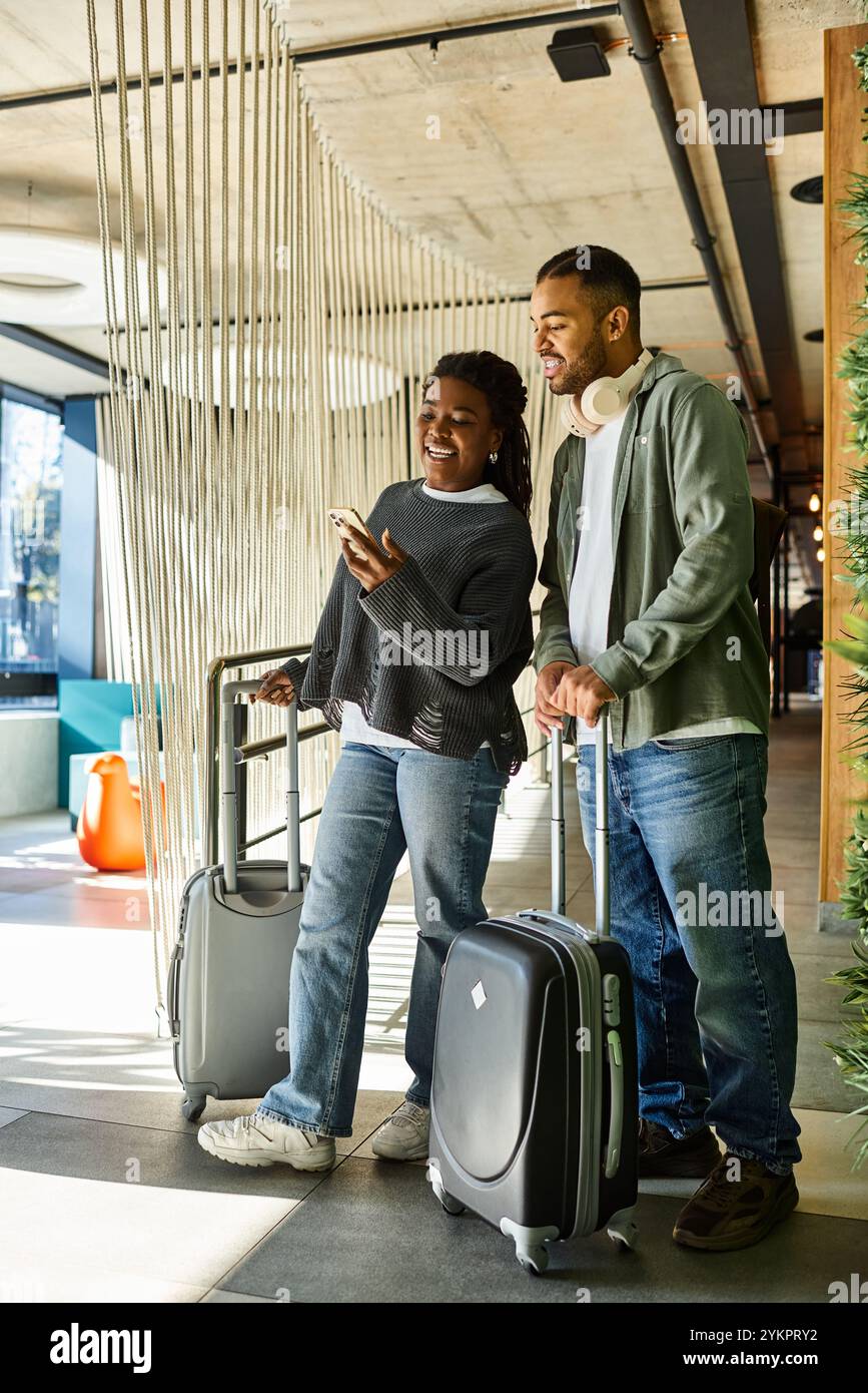 Una giovane coppia con i bagagli sorride mentre guarda un telefono, eccitata per la loro prossima vacanza. Foto Stock