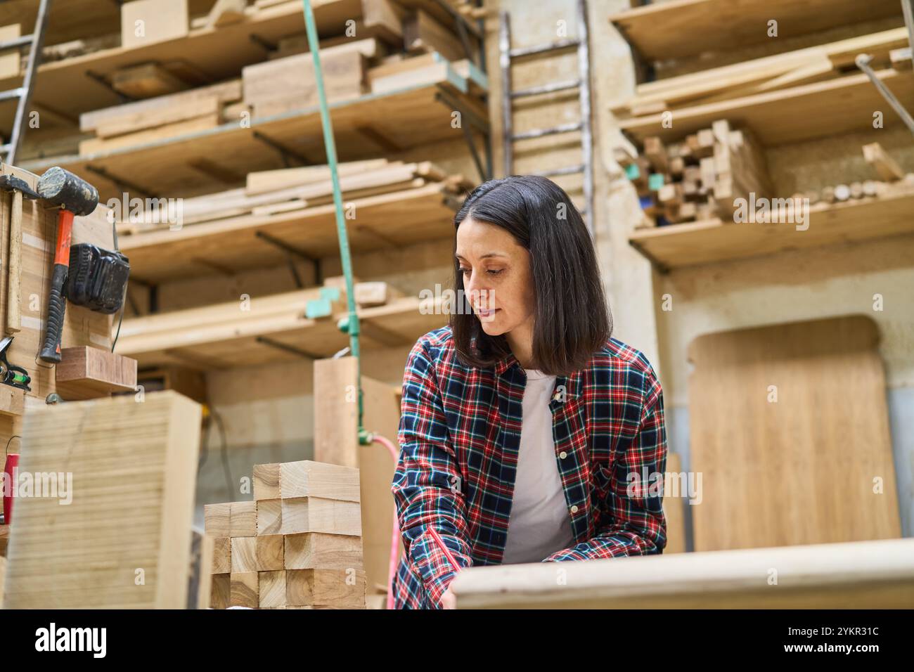 Una giovane donna lavora con cura in un bosco, circondato da legno e attrezzi. La sua attenzione e la sua dedizione mettono in risalto le sue abilità nella lavorazione del legno e nell'artigianato Foto Stock