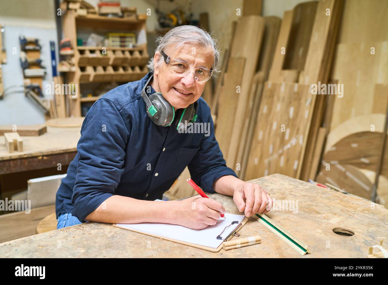 Un esperto artigiano anziano prende appunti in un'officina di legname, concentrandosi sulla precisione e l'esperienza. Circondato da attrezzi e legno, la sua dedizione a c Foto Stock