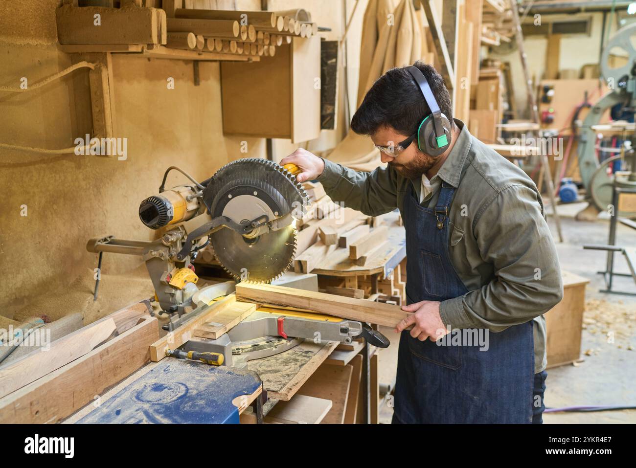 Un lavoratore del legno esperto con protezione uditiva aziona una sega da tavolo in un vivace bosco, circondato da utensili e materie prime, mettendo in evidenza la messa a fuoco e la cr Foto Stock