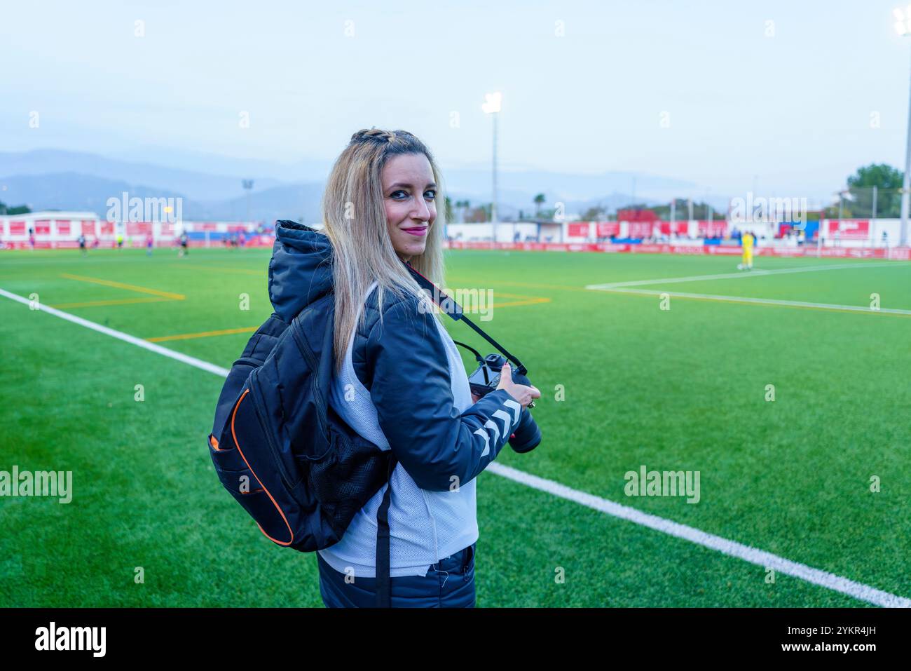 Fotografa sportiva di mezza età sorridente alla fotocamera mentre tiene in mano le attrezzature su un campo da calcio al crepuscolo Foto Stock