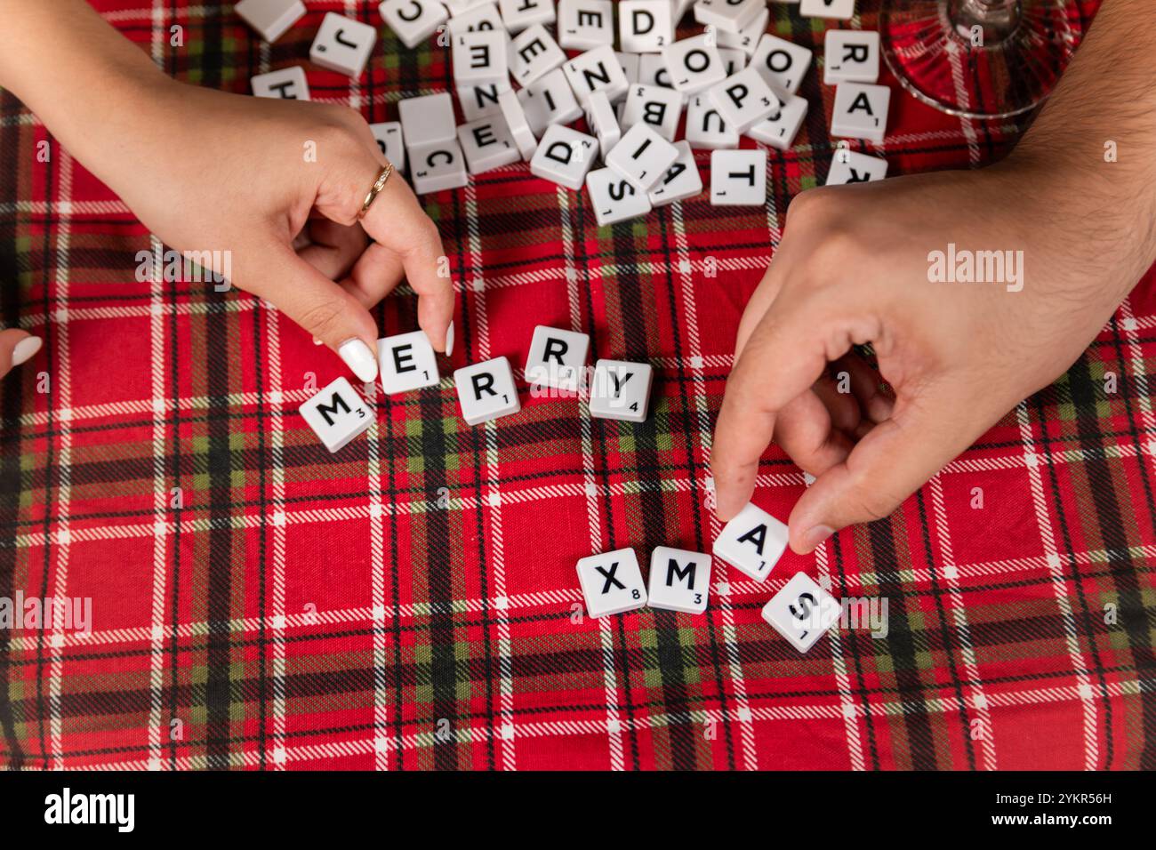Due mani che sistemano le tessere delle lettere per scrivere "Merry Xmas" su una tovaglia a quadri rossa. La scena cattura l'essenza delle vacanze. Foto Stock