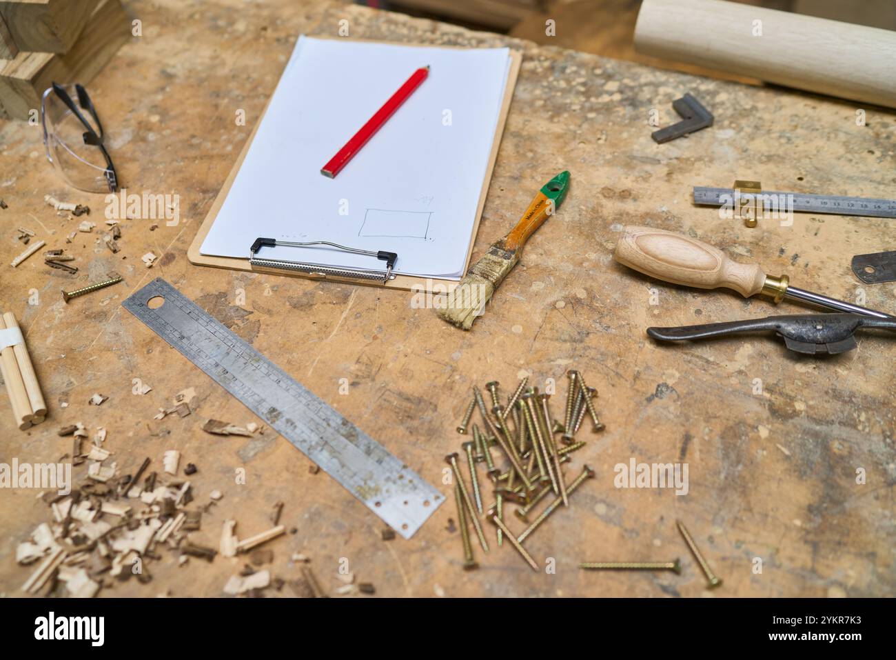 Utensili e materiali assortiti per la lavorazione del legno sono sparsi su un banco da lavoro rustico. La scena include strumenti come un righello, un cacciavite e un pennello, enfatizza Foto Stock