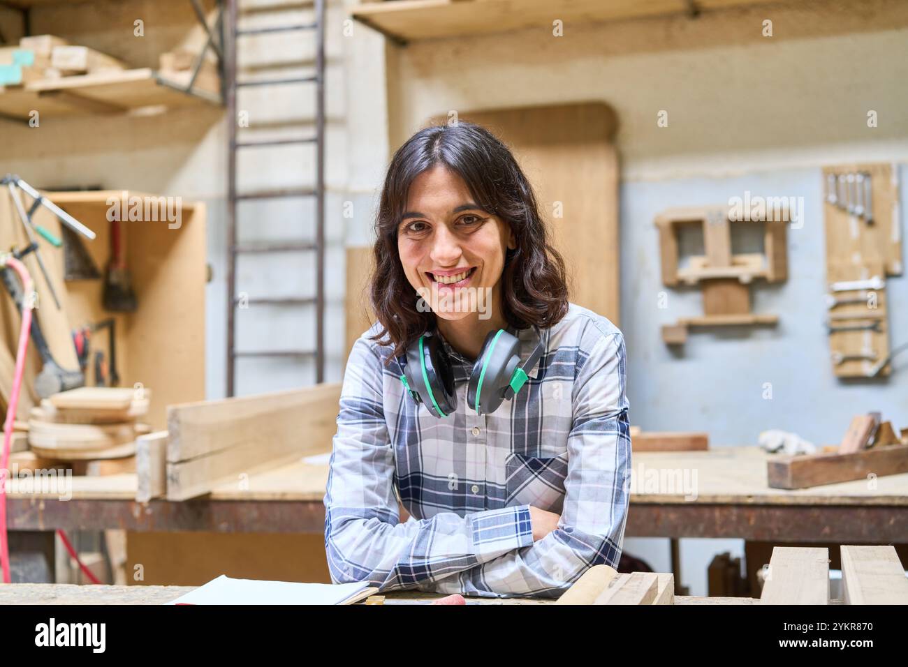 Una giovane donna sorride con fiducia in un laboratorio di lavorazione del legno, mostrando maestria artigianale e creatività. Indossando le cuffie, incarna un approccio moderno Foto Stock