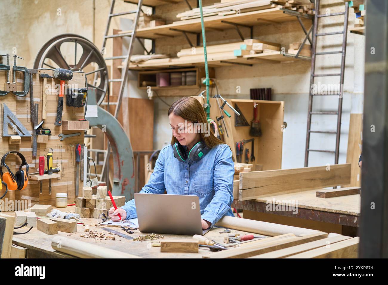 Una giovane donna conduce il lavoro sul suo laptop tra gli utensili per la lavorazione del legno in un bosco. L'impostazione trasmette produttività e maestria artigianale in una b Foto Stock