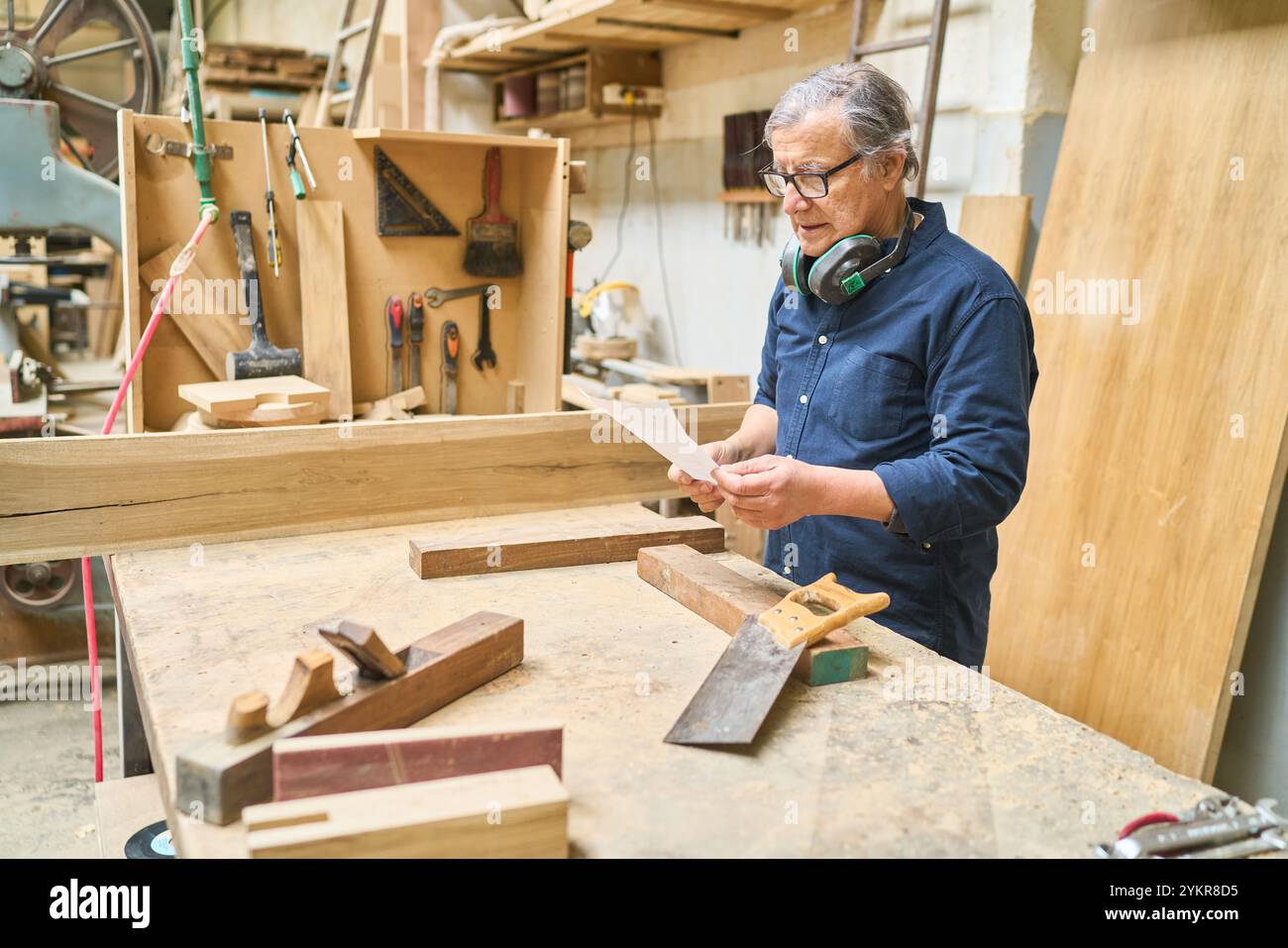 Un artigiano esperto esamina un pezzo di legno in un'officina di legno circondata da attrezzi. L'immagine raffigura la maestria e la dedizione Foto Stock