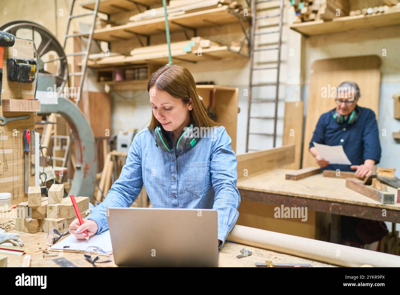 Una giovane donna lavora su un notebook in un bosco, imparando le capacità di lavorazione del legno da un mentore anziano in un ambiente collaborativo. La scena mette in evidenza il tè Foto Stock