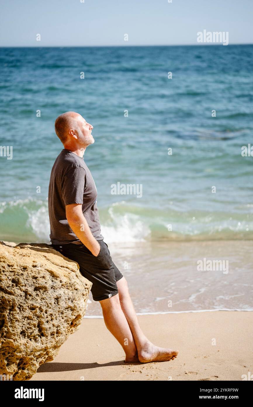 Uomo caucasico seduto su una roccia alla spiaggia di São Rafael ad Albufeira, Algarve - Portogallo Foto Stock