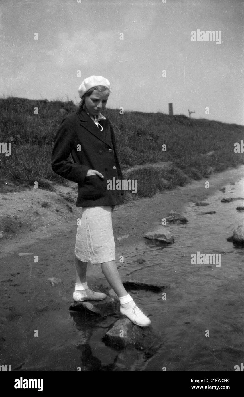 circa1940, ragazza storica e attraente con gonna lunga, blazer, berretto, calzini e scarpe in piedi su rocce in un ruscello. Foto Stock circa1940, ragazza storica e attraente con gonna lunga, blazer, berretto, calzini e scarpe in piedi su rocce in un ruscello. Foto Stock