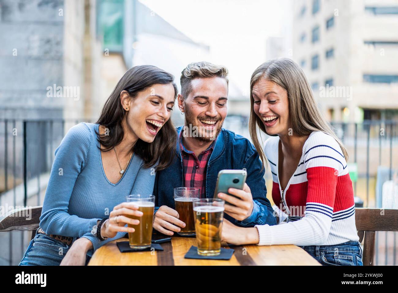 Tre amici che ridono insieme mentre guardano uno smartphone all'aperto. Giovani adulti che si godono un momento di divertimento con una birra in un bar della città Foto Stock