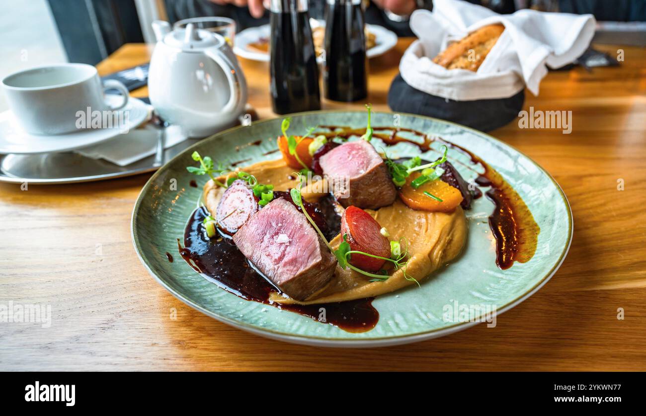 Pezzo di carne d'anatra con barbabietola al forno su purea di castagne con salsa e foglia di pisello germogliata su piatto decorativo sul tavolo del ristorante con utensili. Festival Foto Stock