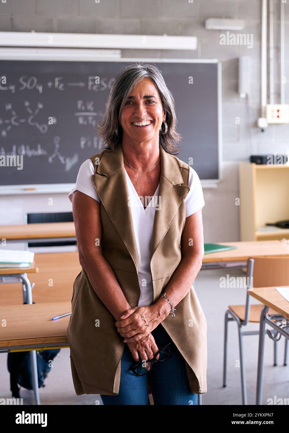 Insegnante di scuola superiore di mezza età che sorride alla macchina fotografica con le mani unite in classe. Foto Stock